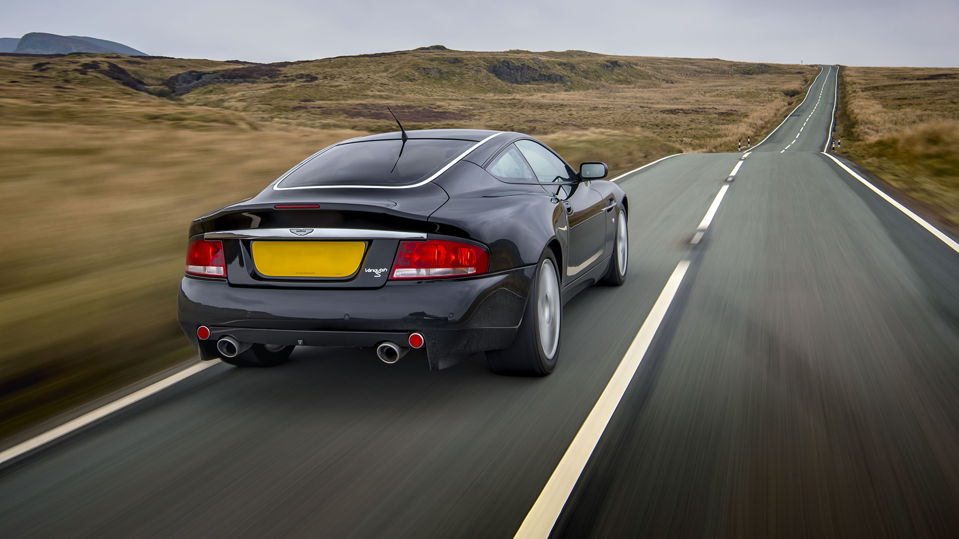 Vue arrière d'une élégante Vanquish S Aston Martin noire naviguant sur une route de campagne pittoresque. 