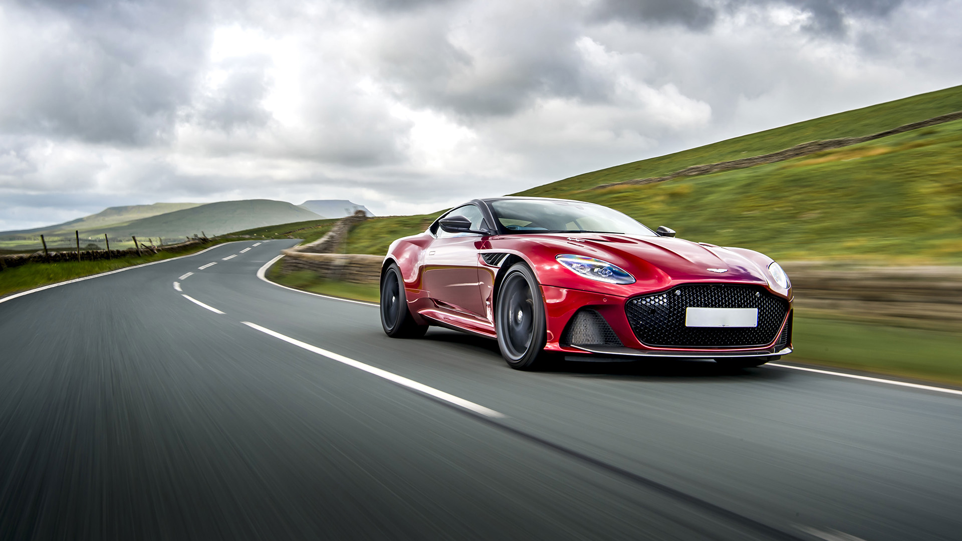 Rouge Aston Martin DBS roulant vite sur une route de campagne avec un ciel gris au-dessus.