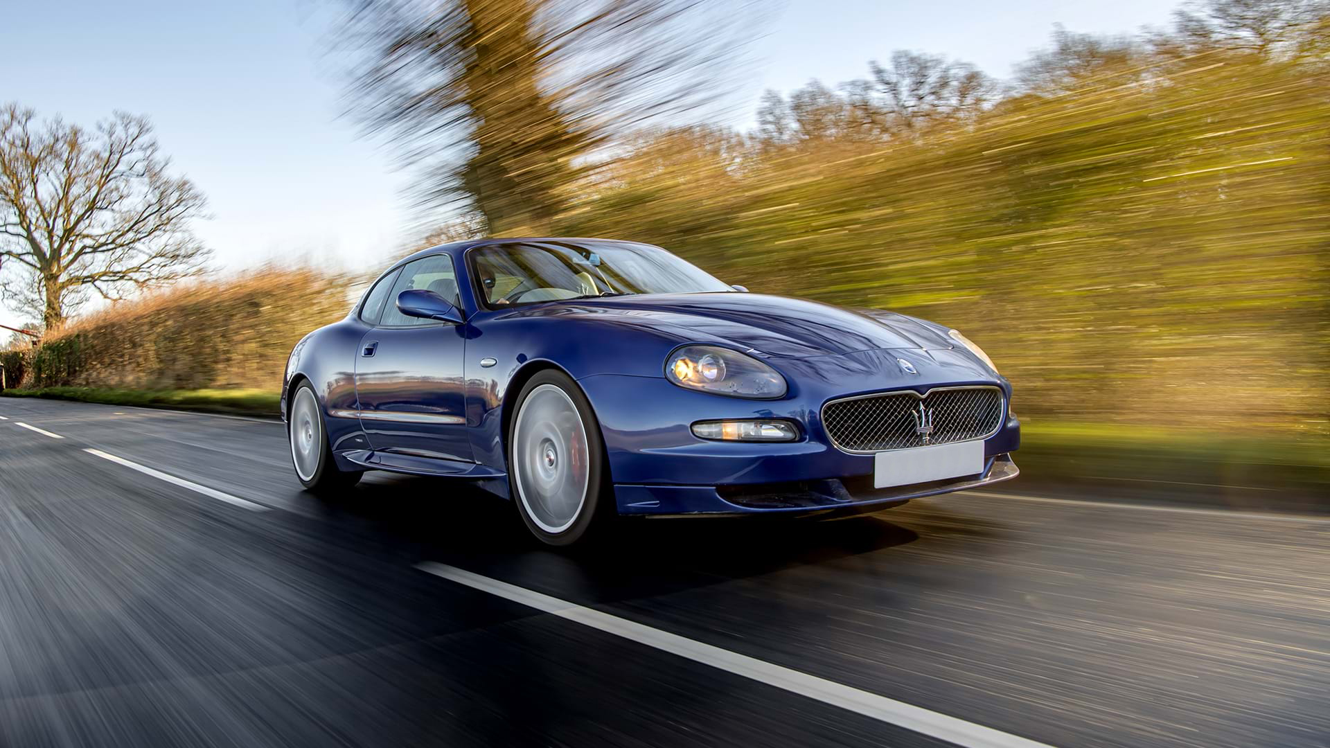 Classic Blue Maserati GranSport conduisant sur une route de campagne avec un ciel bleu au-dessus.