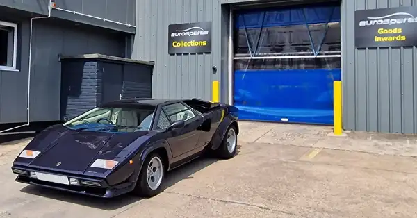 Une Lamborghini Countach bleue est garée devant l'entrepôt d'Eurospares, avec des panneaux « enlèvement par le client » et « réception des marchandises » affichés au mur.